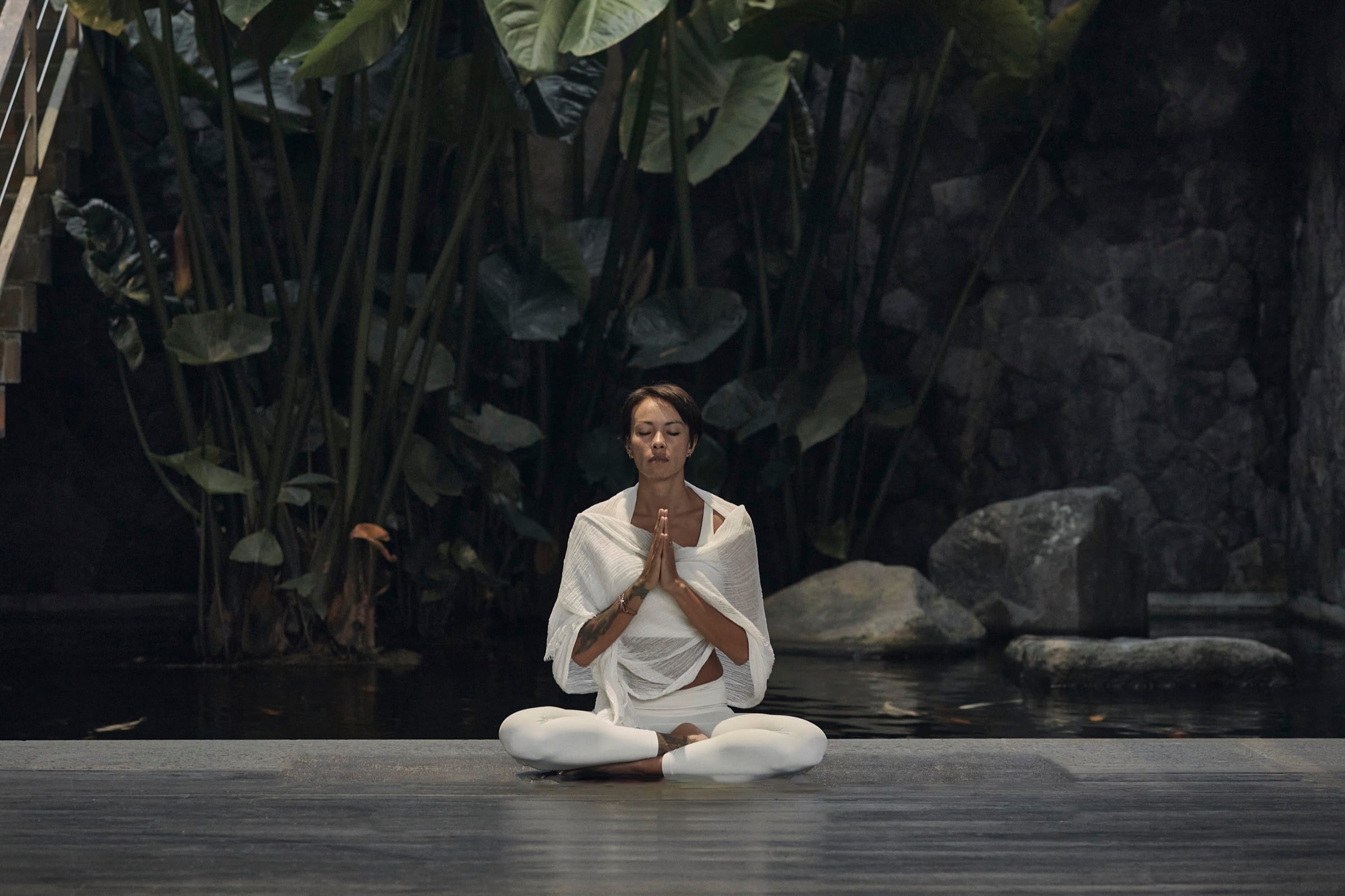 Kimberley Utama sitting cross legged beside a still pond in Ubud, hands at her heart, wrapped in a soft white shawl with tropical leaves rising behind her.
