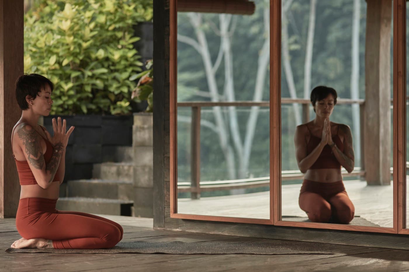 Kimberley kneeling with her hands at her heart, her reflection held in a tall mirror that looks out over the jungle.