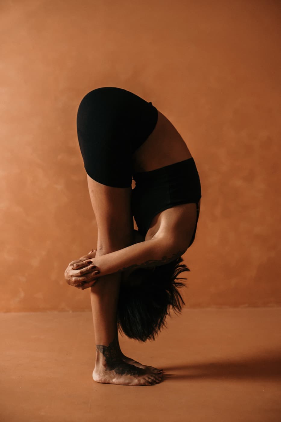 Kimberley folding forward with hands clasped behind her calves, standing against a warm terracotta wall.