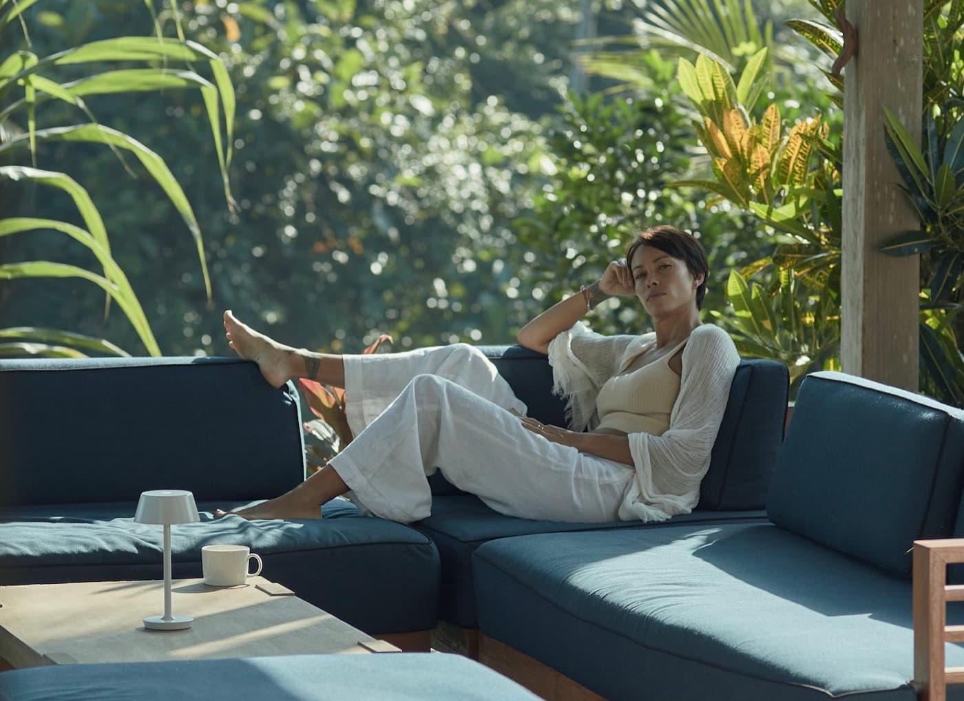 Kimberley resting on an outdoor sofa with a cup of tea beside her, jungle greenery filling the view behind.