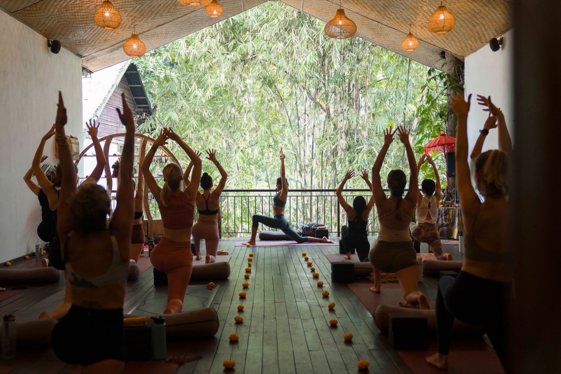 A group of trainees kneeling with arms lifted in a morning practice on an open air deck, marigolds lining the floor, with Kimberley leading from the front in a low lunge.