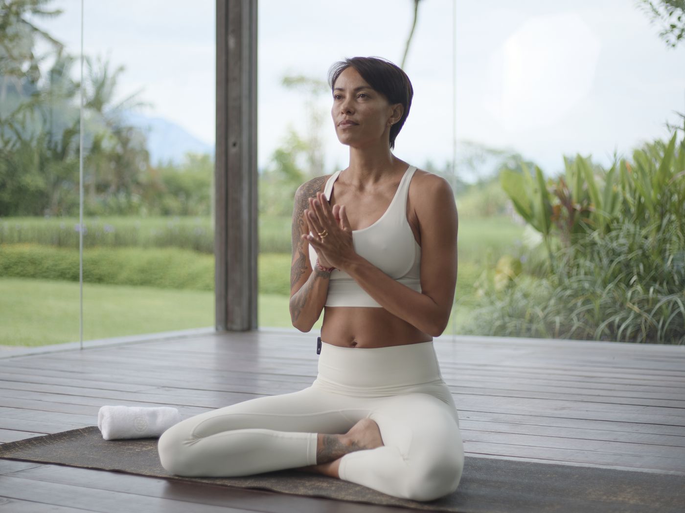 Kimberley seated on a yoga mat outdoors, hands at heart, wearing a headset microphone