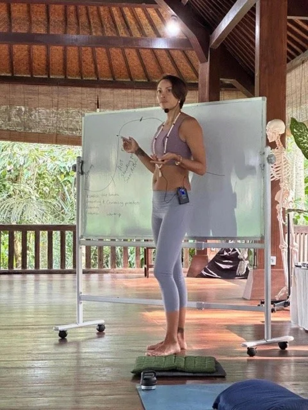 Kimberley teaching anatomy at a whiteboard beside a skeleton model, in an open air shala.