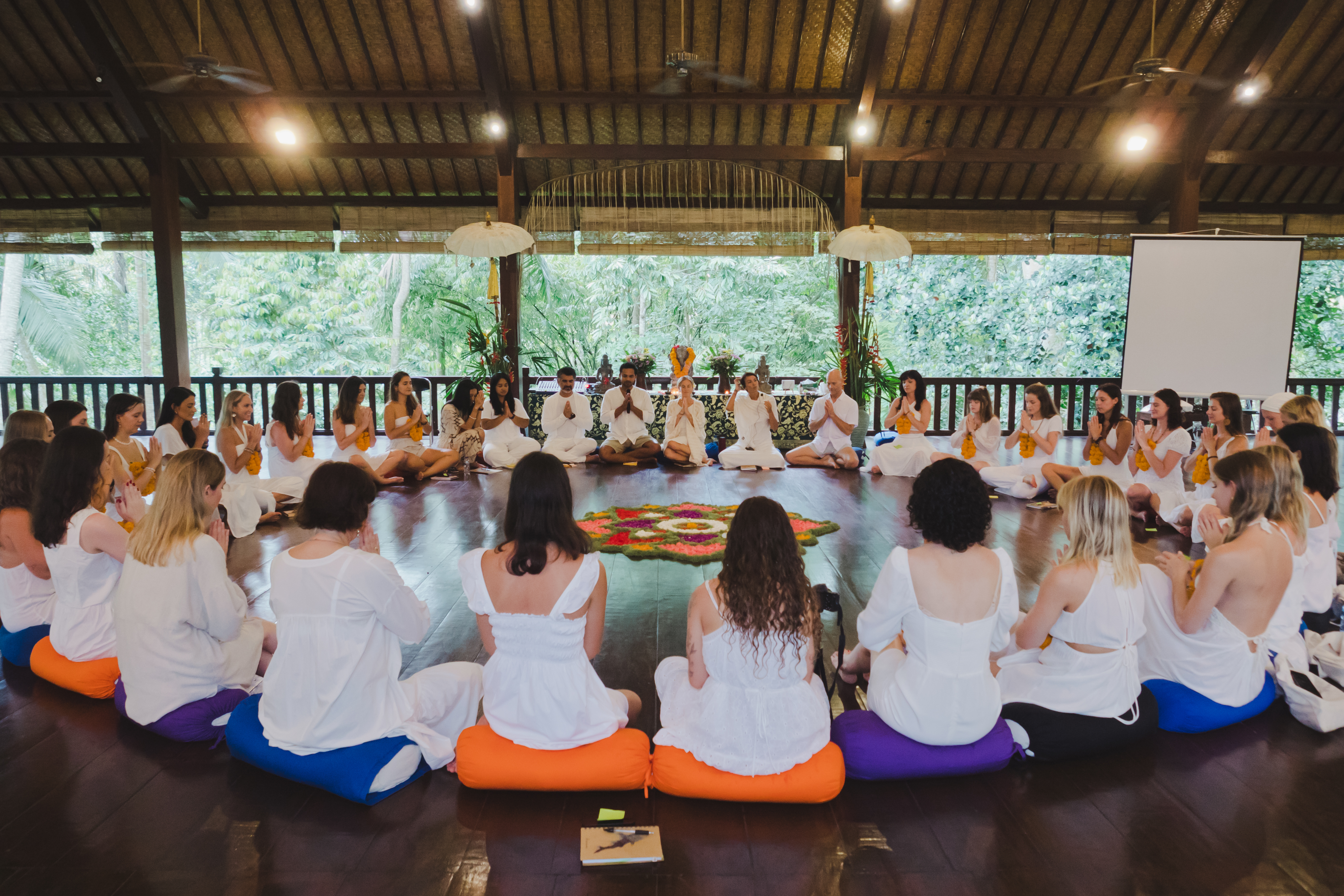 Trainees dressed in white sitting in a full circle with hands in prayer around a flower mandala during a training ceremony